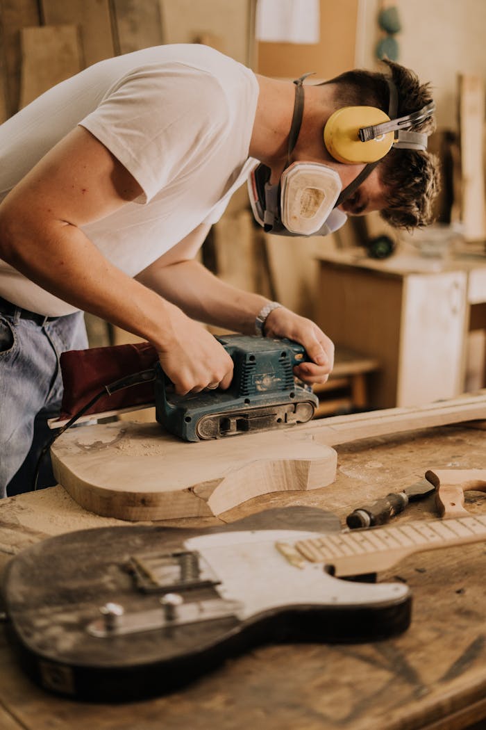 Man sanding guitar body in a workshop using a sander, surrounded by woodworking tools.